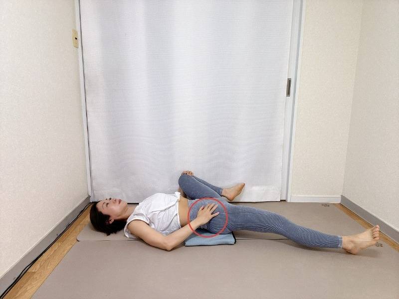 お尻の下にブランケットを敷いた仰向けから左脚の付け根に手を添えて膝を伸ばし、右膝を胸に寄せて外に開いた内転筋のストレッチ