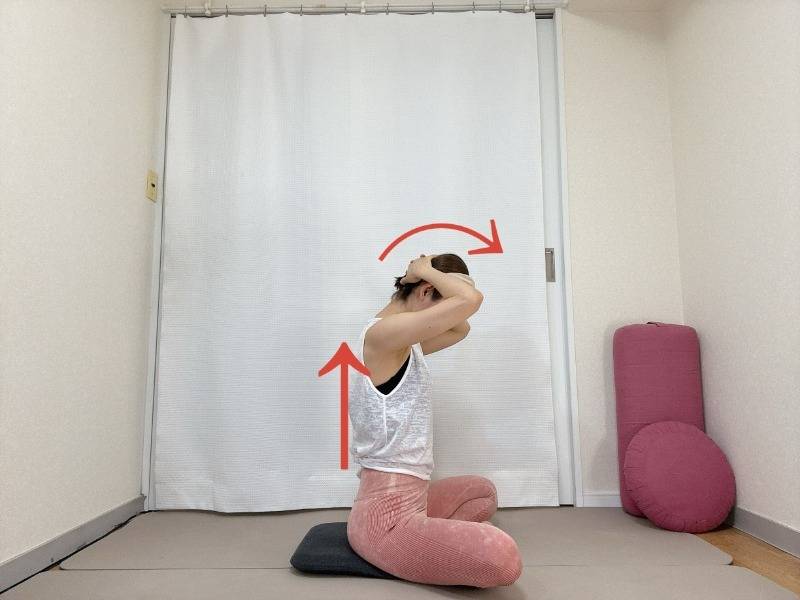 あぐらで座り、頭の後ろで両手を組み、背筋を伸ばして頭を前に倒す首の後ろのストレッチ