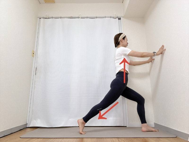 両手を壁につき、右膝を曲げ、左脚を後ろに引いて脚の付け根を伸ばす腸腰筋のストレッチ
