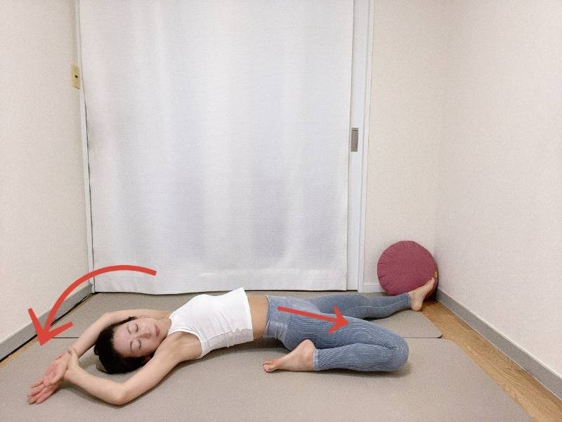 左膝を曲げた半分の割座から仰向けになり両手を頭上に伸ばし、右手で左手首をつかんで上体を左に傾けたストレッチ