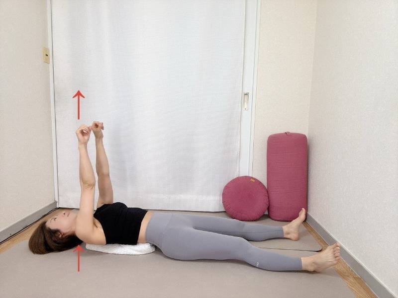 丸めたバスタオルを肩甲骨の下にセットした仰向けから肩甲骨を浮かせて両手を天井に伸ばした体勢