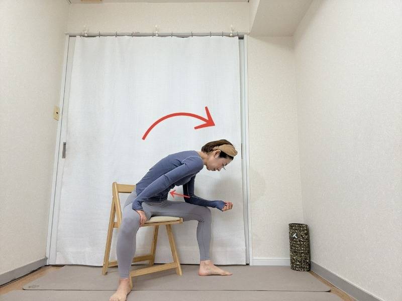 脚を大きく開いて椅子に座り、右肘を腿に乗せて背中を丸めて上体を右斜め前に倒す腰痛予防ストレッチ