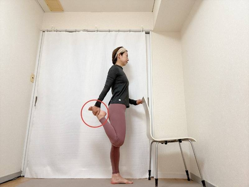 椅子の背もたれに右手を添え、左膝を曲げて脚の甲を掴んで立った体勢