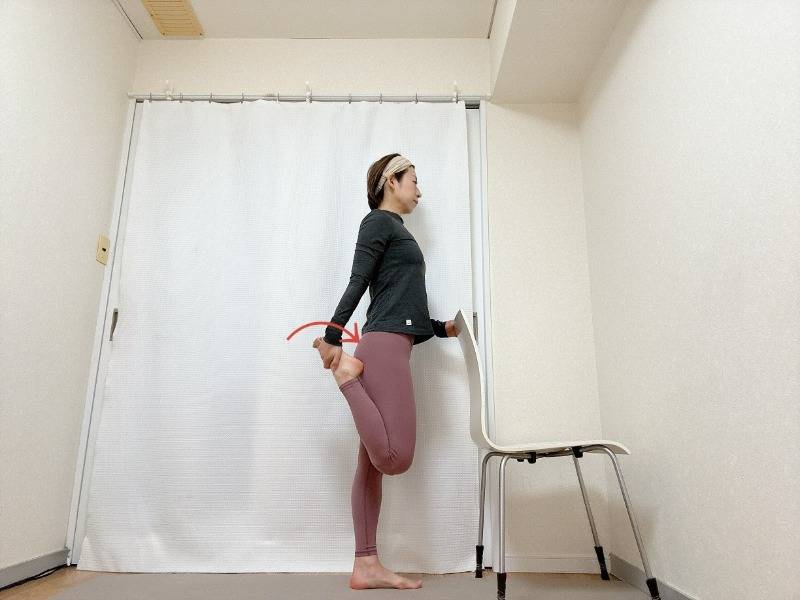 椅子の背もたれに右手を添えて立ち、左膝を曲げて脚の甲を掴んでかかとをお尻に引き寄せた体勢