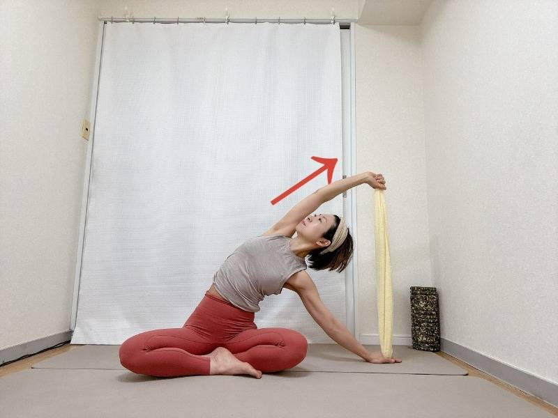 あぐらでタオルの両端を持ち、右手を床に下し、左肘を伸ばして上体を右に傾け、左わき腹を伸ばすストレッチ