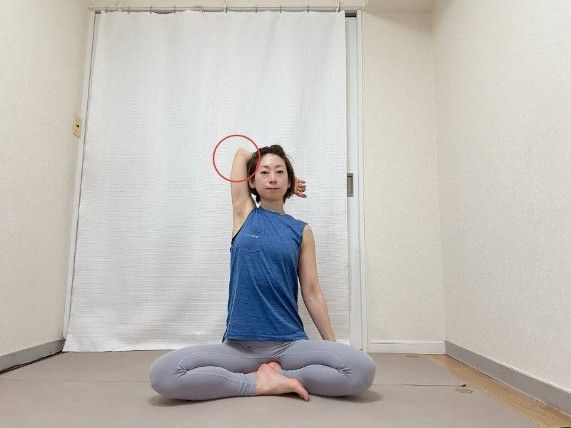 左肘を曲げて高く引き上げ、腕を耳の横に近づけて脇を大きく開いてあぐらで座った体勢