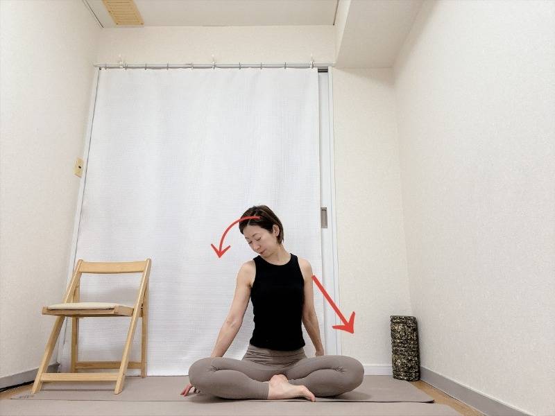 右腕を斜め後ろの床へ伸ばしたあぐらで座り、頭を左斜め下へ傾け、右の首筋から肩まわりを伸ばすストレッチ