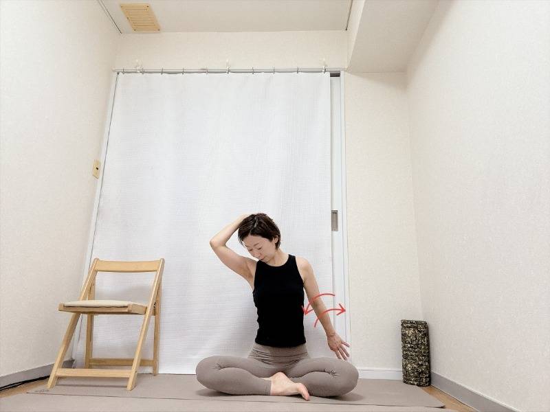 あぐらで座り、左手を後頭部に添えて頭を左下に傾け、後ろに引いた右腕後ろに引いた右腕を前後にひねる肩まわりのストレッチ
