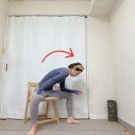 脚を大きく開いて椅子に座り、右肘を腿に乗せて背中を丸めて上体を右斜め前に倒す腰痛予防ストレッチ