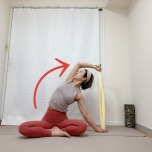 あぐらでタオルの両端を持ち、右手を床に下して左肘を曲げ、上体を右に傾けて左わき腹を伸ばすストレッチ
