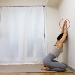 壁の前に正座で座り、両手を重ね、手のひらを頭上の壁につき、脇と胸を開くストレッチ