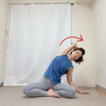 左肘を曲げて高く引き上げ、腕を耳の横に近づけて脇を大きく開いてあぐらで座り、上体を右に傾けた体勢